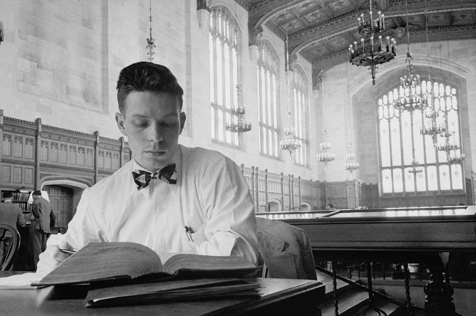 University of Michigan male student studies in a gothic reading room with arched windows and chandeliers. He is caucasian and wears a bow tie. (Image credit: Stanley Kubrick, Look magazine photograph collection, Library of Congress.)