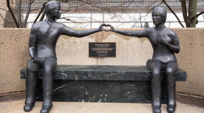 Sculpture at Michigan Medicine facility in Ann Arbor features two health care workers on opposite ends of a bench, reaching toward one another and creating a heart with their respective hands.