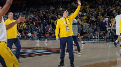 Kim Barnes Arico's son, Trevor Arico, was part of the University of Michigan men's staff as he began a career in coaching. Image shows Arico in maize sweatshirt and blue pants with arm raised at center court.
