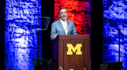 University of Michigan president Domenico Grasso stands at podium with Block M in front of blue and red panels.