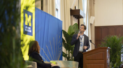Man with microphone presents from podium during AI summit, flanked by maize and blue drapes and Block M signage.