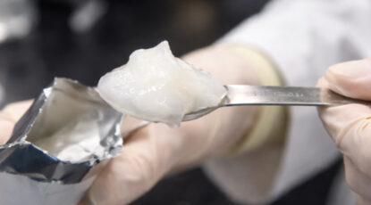 Close-up of scientist's hand in rubber glove holding a spoon containing a shiny white gel.