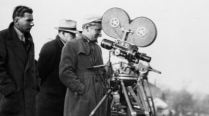 Black and white image of three men behind a movie camera, circa 1930.