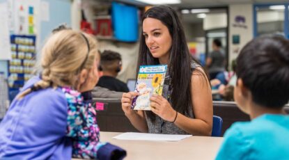 Teacher holds book up for young child in what appears to be a library setting.