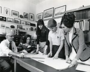 Black and white image of four women standing at a table looking at documents while male professor is seated. U-M, Ann Arbor. 1974.
