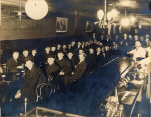Sepia-toned bar scene of a long table with men in commencement caps and gowns drinking pits of beer. University of Michigan somewhere between 1913-18.