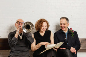 A woman sits between two men holding a book. One man plays a trombone, the other wears a red corsage. They are founders of the Bang On a Can musical collective.
