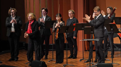 Musicians dressed in formal wear stand on stage at the University of Michigan's Rackham Auditorium and applaud composer Julia Wolfe.