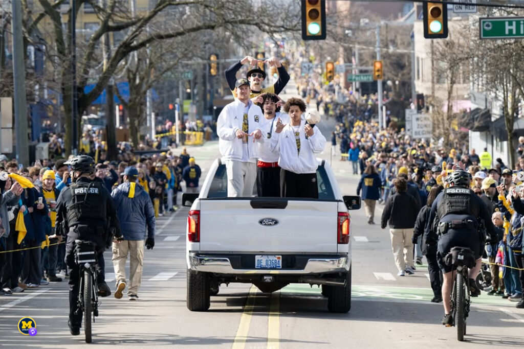 Members of the University of Michigan men's basketball team strand in the flatbed of a white pickup truck traveling down State Street as they celebrate their 2026 NCAA championship victory.