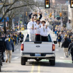 Members of the University of Michigan men's basketball team strand in the flatbed of a white pickup truck traveling down State Street as they celebrate their 2026 NCAA championship victory.