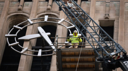 Worker in hardhat and dayglo hoodie stands on scaffolding outside the top of Burton Memorial Tower at University of Michigan in Ann Arbor; next to the face of the clock itself with a crane in the foreground.