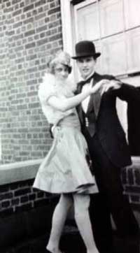 A black and white photo of a couple wearing formal dress of the 1920s as if preparing to go dancing.