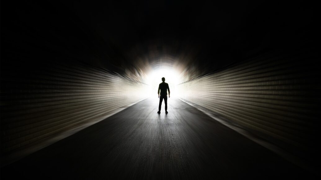 Lone figure stands in a dark tunnel with white light at the end.