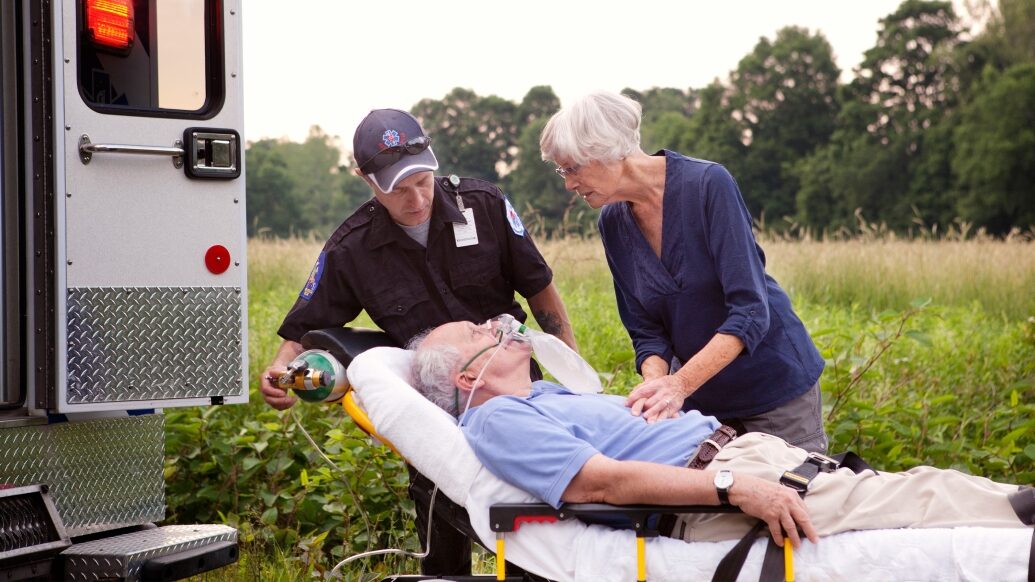 Elderly patient lays on a gurney while elderly companion comforts him and EMT loads him into waiting ambulance.