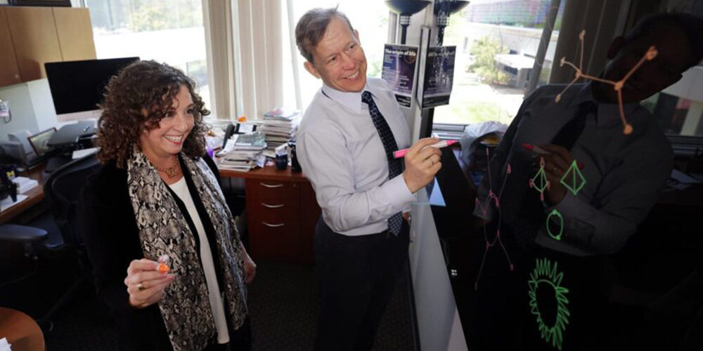 Two professors hold markers and write on a board. Man and woman, caucasian.