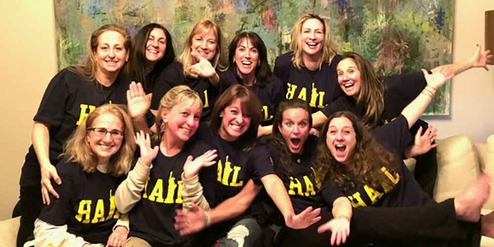 A pack of Michigan alumni, caucasian females dressed in Hail shirts, looks very happy.