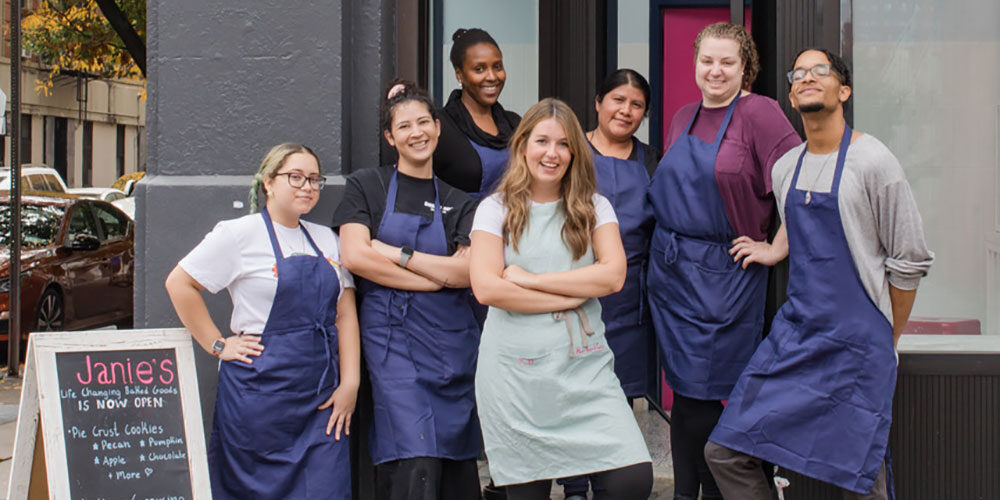 Janie Deegan and her bakery staff stand together in front of the shop, dressed in aprons with a Janie's sign next to them.