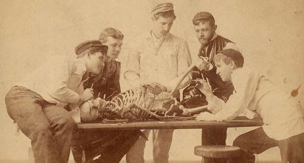 Five young men crowd around a table, on which a skeleton lies. The men are medical students of the late 19th/early 20th century. The image is sepia. A youngish man in cap gestures with a pencil while the others observe. An early anatomy ab at the University of Michigan.