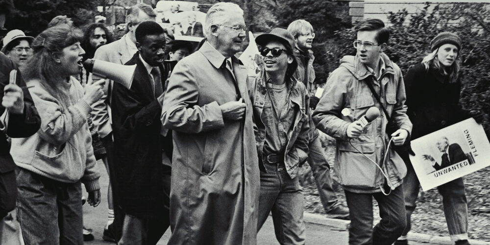 Older man in white hair and trenchcoat surrounded by protesters with bullhorns and signs. U-M campus, 1988. Robben Fleming.