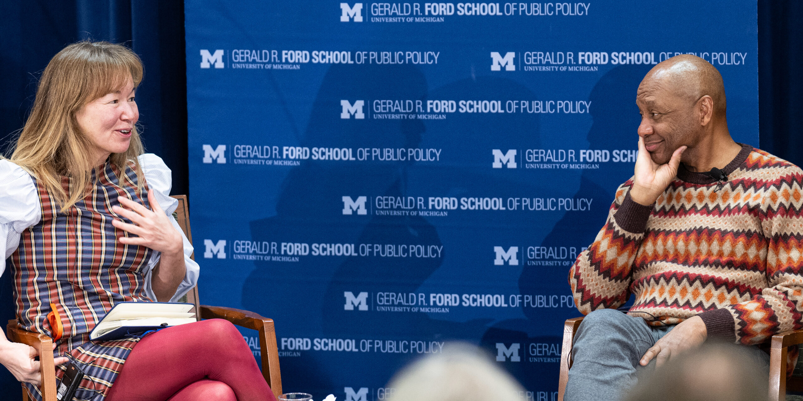 A man and woman sit on stage for an interview in front of a blue banner with white lettering that reads Gerald R. Ford School of Public Policy;