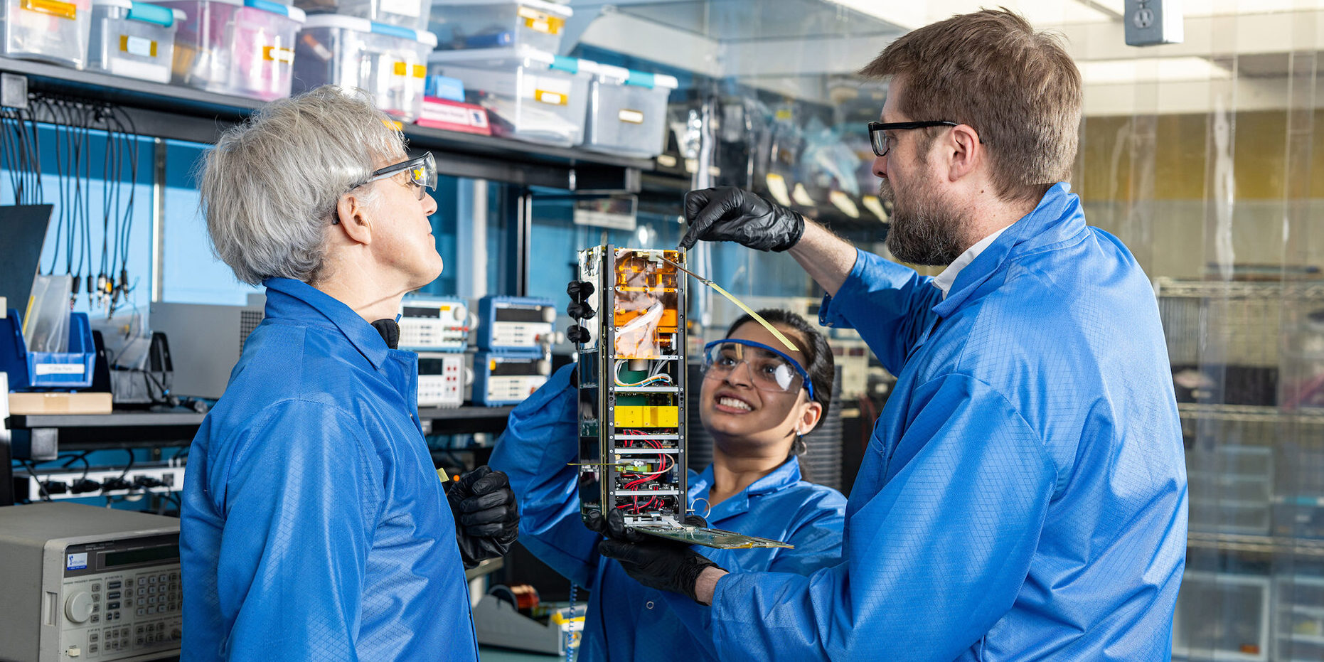 Scientists observe some kind of machine in a lab. They are wearing blue coats and protective glasses and are surrounded by machinery.