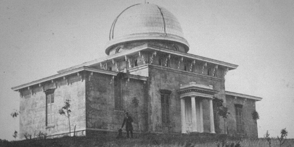 19th century b&w photo of a man in front of the domed Detroit Observatory at U-M in Ann Arbor. The man in the photograph is thought by some to be Henry Tappan himself, with one of his dogs, Leo or Buff.