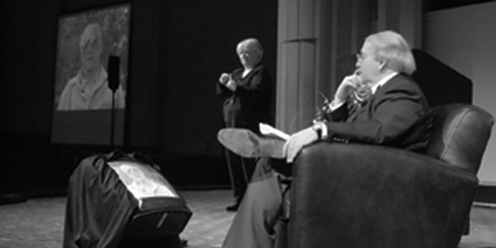 Black and white image of caucasian man in easy chair on a stage interviewing a person on a giant video monitor. The people are Enoch Brater, University of Michigan professor emeritus, and playwright/alumnus Arthur Miller.