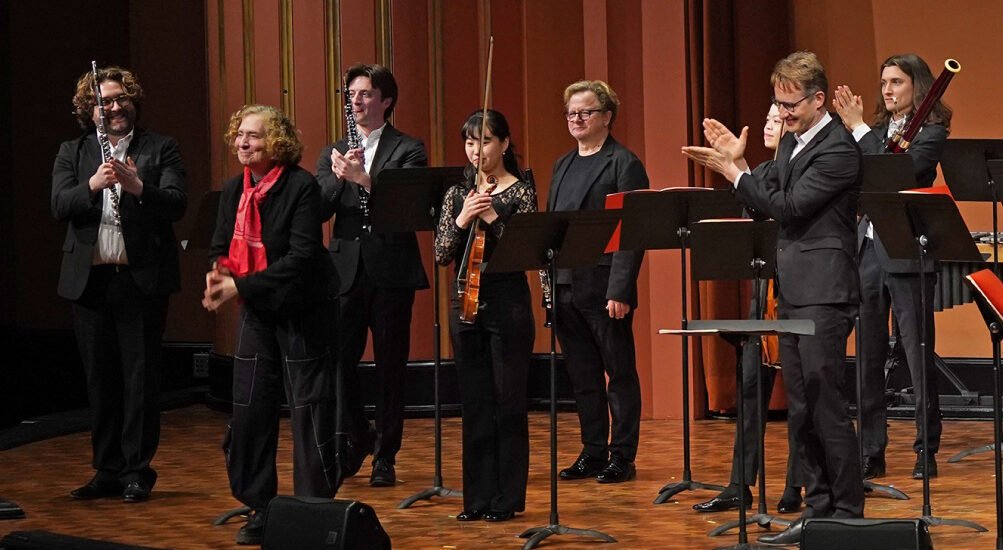 Musicians dressed in formal wear stand on stage at the University of Michigan's Rackham Auditorium and applaud composer Julia Wolfe.