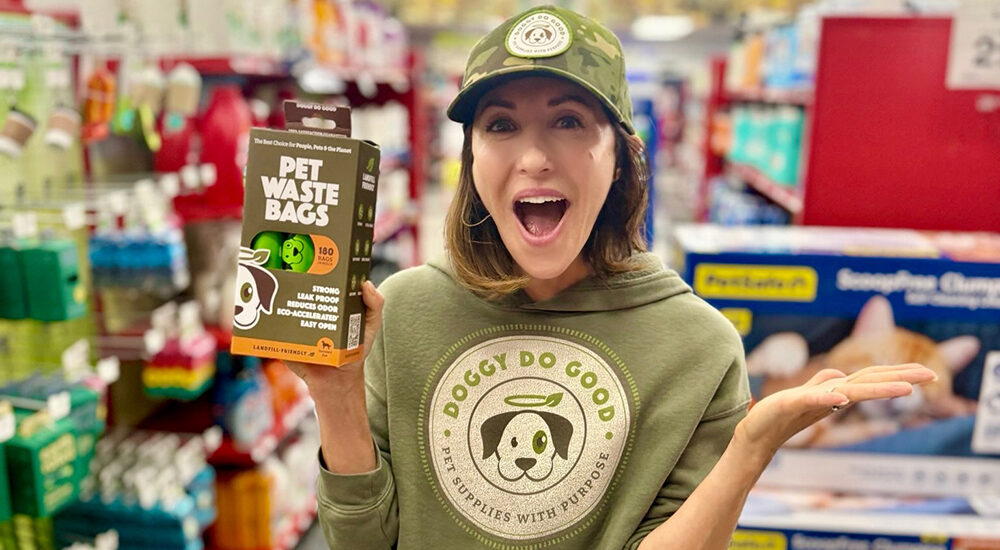 Cara AnnMarie, a caucasian actress and founder of Doggy Do Good stands in pet store dressed in branded gear holding a package of pet waste bags. She is a graduate of U-M in Ann Arbor.